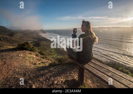 A blonde hiker rests on a bench that overlooks the pacific ocean in a beautiful portion of the California coastline in Point Reyes national seashore. Stock Photo