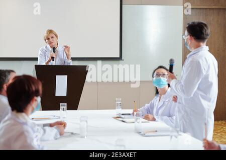 Serious female speaker with vial in hand listening to question from the audience when standing at tribune at medical conference Stock Photo