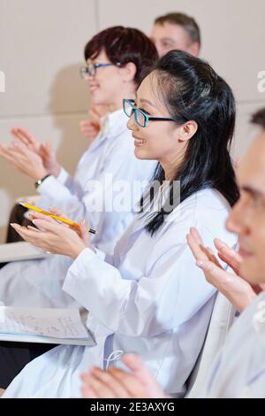 Young smiling scientists in labcoats applauding to speaker at medical conference Stock Photo