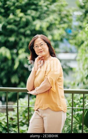 Portrait of cheerful senior Vietnamese woman in glasses standing outdoors on balcony and smiling at camera Stock Photo