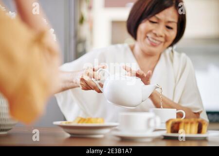 Senior Asian woman sitting at kitchen table and pouring hot tea in cup, selective focus Stock Photo