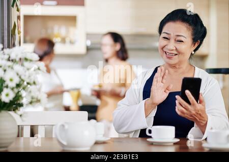 Portrait of happy senior Vietnamese woman waving with hand when video calling her friends or family members Stock Photo