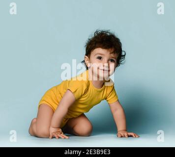 Small cute positive curly baby boy toddler in yellow comfortable jumpsuit crawling on floor and looking at camera Stock Photo