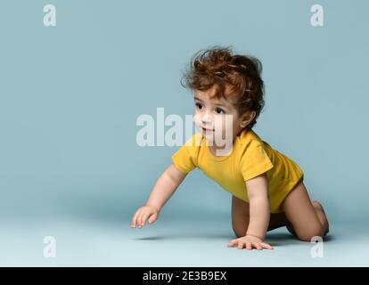 Cute smiling curly baby boy toddler in yellow comfortable jumpsuit crawling on floor and feeling cheerful Stock Photo