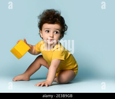 Small cute smiling curly hairy baby boy toddler in yellow comfortable jumpsuit sitting on floor with cube toy and looking aside Stock Photo