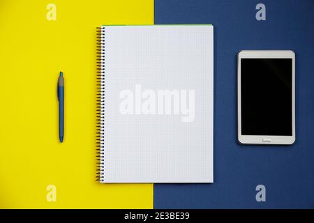 Top view of an open notebook, a tablet pc, and a pen against blue and yellow background Stock Photo