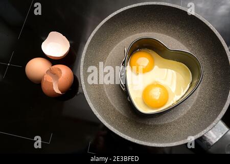 Two fried eggs in heart shape on a frying pan and empty shells on the black stove, cooking with love for a valentines day breakfast, copy space, high Stock Photo