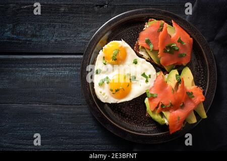Two sandwiches with avocado and smoked salmon and two fried eggs in one heart shape, romantic valentines day meal for a couple on a dark blue wooden t Stock Photo