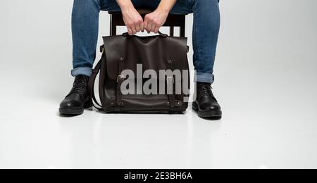 Man in a blue jeans and black boots sits on a chair with a brown men's shoulder leather bag for a documents and laptop on a white floor. Mens leather Stock Photo