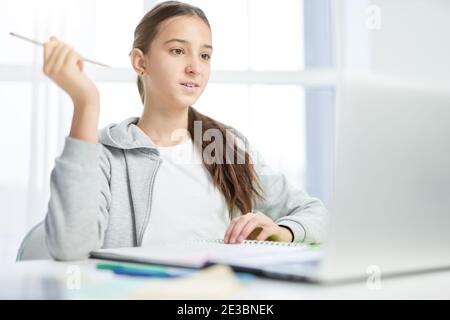 Pretty latin teenage girl looking at the screen of laptop while communicating with her teacher, having online lesson at home. Distance education, home schooling concept Stock Photo