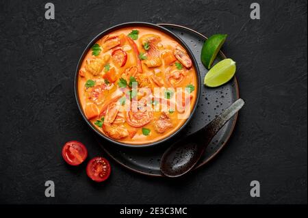 Moqueca with Fish and Shrimps in black bowl on dark slate table top. Brazilian sea food curry dish with coconut milk and vegetables. Top view Stock Photo