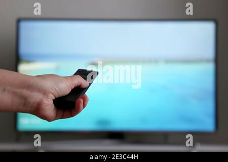 Female hand with remote control on TV screen background. Woman switches channels, watching travel programs, planning vacation during the coronavirus Stock Photo