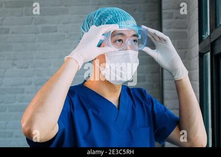 Male medical worker putting on protective gears like latex gloves, surgical face mask, bouffant cap, medical goggle at work. Asian man wearing and getting ready for working at hospital Stock Photo