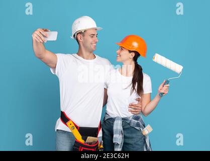 Cheerful Repair Workers Couple In Protective Hard Hats Taking Selfie On Smartphone Stock Photo