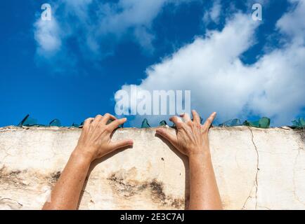 Hands on broken glass on top of wall with blue sky copy space. Escaping, trapped, prison, lockdown... concept.