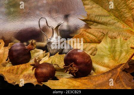 Still life with autumn dry sycamore leaves, dry pomegranates and decorative jug close-up on blurred background