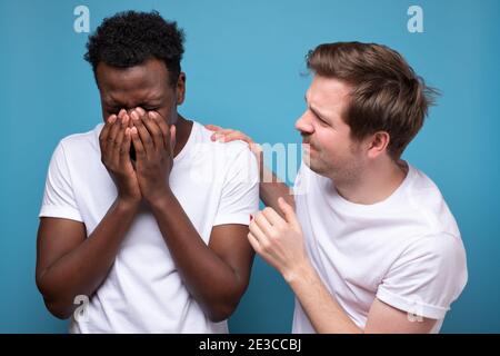 Caucasian man calms down his african colleague or friend, who has troubles, miserable facial expressions, keeps hands on his shoulders, stands closely Stock Photo