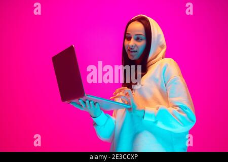 Holding laptop, working. Caucasian woman's portrait on pink studio background in mixed neon light. Student, worker. Concept of human emotions, facial expression, sales, ad, fashion. Copyspace. Stock Photo