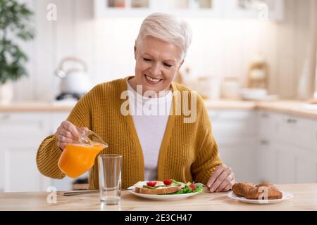 Attractive old lady pouring juice, having lunch in kitchen Stock Photo