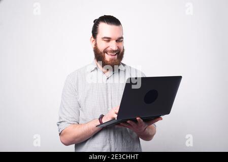 Portrait of happy bearded man in shirt using laptop over white wall.