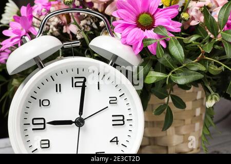 White alarm clock against the background of fresh flowers in a wicker basket. Spring time. Stock Photo