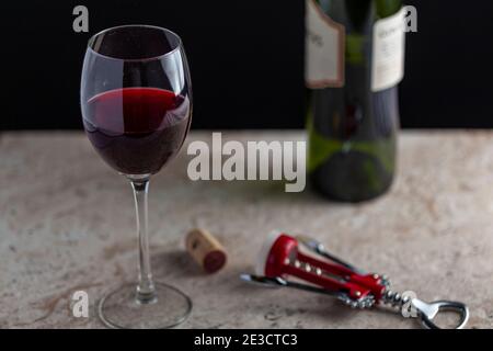 Image of a standard size wine glass with red wine filled halfway through. There is a corkscrew on the marble countertop with a cork lying beside. A gr Stock Photo