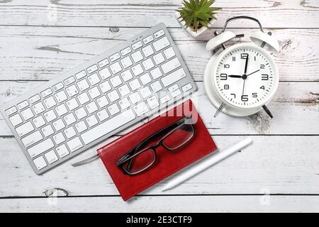 Flat lay view of a red notebook, white keyboard, pen and black glasses on a white wooden background. Top view of modern office workplace. Stock Photo