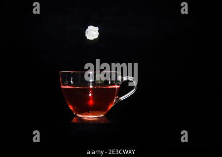 A piece of rock sugar is splashing into a glass cup of tea in front of a black background Stock Photo