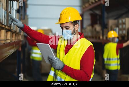Young man working in warehouse doing inventory using digital tablet and loading delivery boxes plan while wearing face mask during corona virus Stock Photo