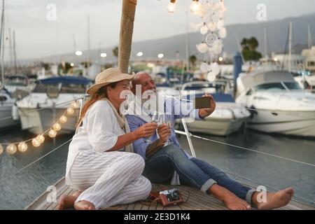 Senior couple toasting champagne while taking selfie on sailboat vacation - Happy mature people having fun celebrating wedding anniversary Stock Photo