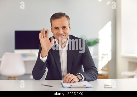 Smiling businessman sitting at office desk and waving hand at camera saying hello during video call Stock Photo