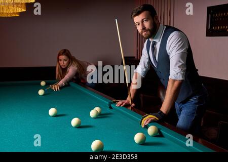 young active friends are playing billiards in bar after work, have rest and leaisure time, preparing aiming to shoot pool balls Stock Photo