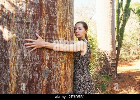 Young woman spreading her arms around huge baobab tree trunk, hugging it, eyes closed Stock Photo