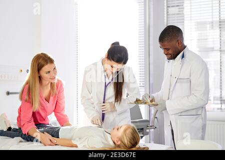 team of doctors working together while treating kid girl patient lying on hospital bed, african man and caucasian woman consulting girl and holding me Stock Photo