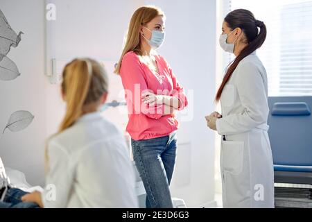 sick girl sits looking at mother talking with doctor in hospital, waiting for solution, how to treat, in medical masks. coronavirus, covid-19. focus o Stock Photo