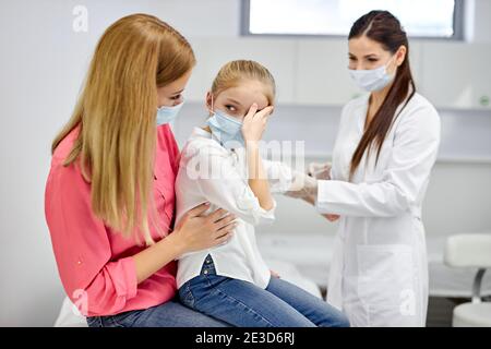 female doctor taking blood sample test of a little girl in the clinic, scared girl sits with mother, woman technician making blood tests on child in t Stock Photo