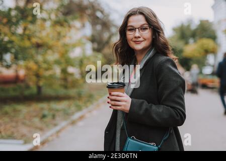 Gladsome bespectacled young lady holding a paper cup of coffee and smiling while standing in the street Stock Photo