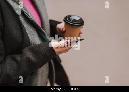 Cropped photo of a lady outdoors having a paper cup of coffee to go and a modern smartphone in her hands Stock Photo