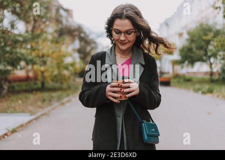 Pretty curly young woman smiling and looking at a paper cup of coffee in her hands while standing in the street Stock Photo