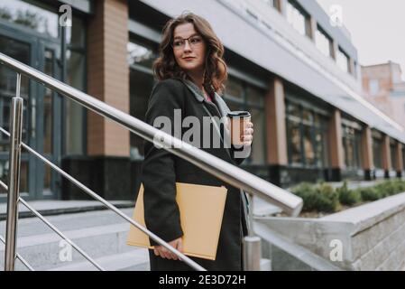 Calm pretty lady looking into the distance with a slight smile while walking downstairs and having a cup of coffee Stock Photo