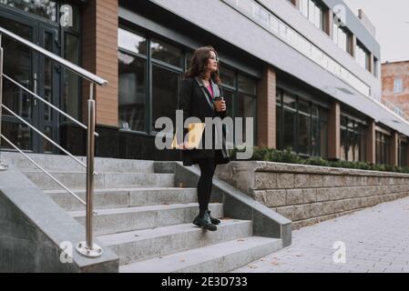 Pretty bespectacled young female having coffee outdoors and looking away while standing on the stairs with a book Stock Photo