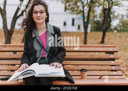 Smiling female student sitting on the bench with hot drink and book outdoors Stock Photo