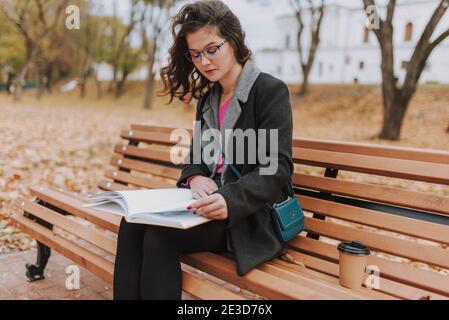 Attractive young lady wearing coat and glasses reading book while sitting on bench outdoors Stock Photo