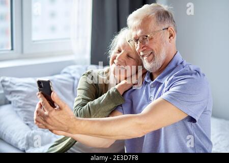 elderly couple taking photo on smartphone, while sitting in bedroom, sit smiling. senior people society lifestyle technology concept. man and woman sh Stock Photo