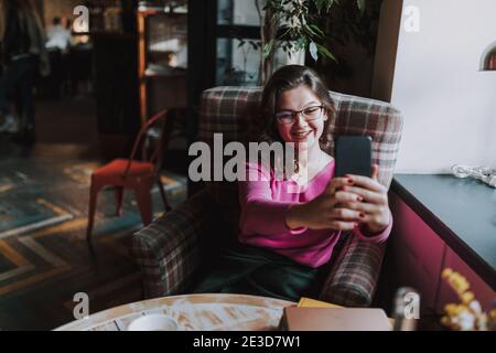 Happy young lady sitting at the table in cafe while taking selfie on smartphone Stock Photo