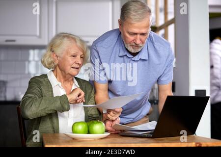 elderly man and woman managing family monthly budget together, focused married couple using computer banking application, counting bills in the kitche Stock Photo