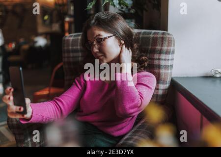 Smiling pretty young woman holding smartphone and taking selfie while resting in coffee house Stock Photo