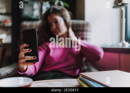 Smiling beautiful young lady holding mobile phone and making selfie while resting in cafe Stock Photo