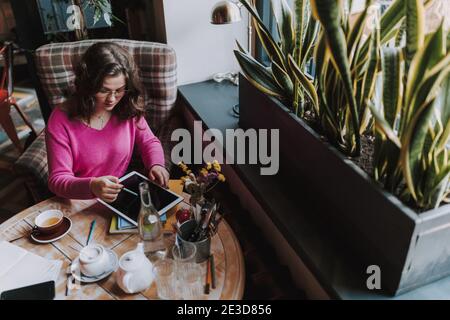 Top view of young lady resting in coffee house and using digital tablet while looking at screen Stock Photo