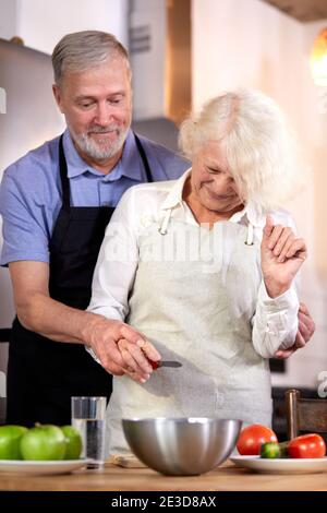 elderly couple preparing vegetable salad in kitchen, gray-haired handsome man helps wife with cooking, going to have healthy breakfast Stock Photo